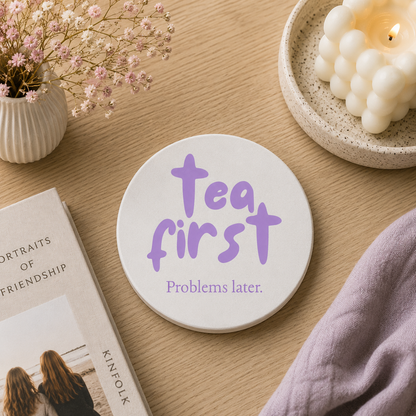 Round ceramic coaster featuring the humorous sarcastic quote "Tea first, problems later." in crisp purple lettering. The coaster is on a table with books and a candle.