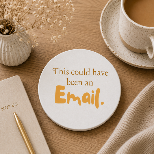 Round ceramic coaster featuring the humorous sarcastic quote "This could have been an email" in sunny yellow lettering. The coaster is on a wooden table with a cup of tea and notebook.