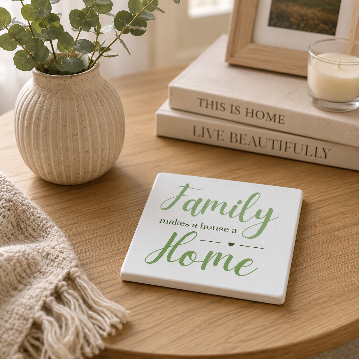Square ceramic coaster featuring the meaningful quote "Family makes a house a home" in elegant green lettering. The coaster is on a wooden side table with books and photo frames.