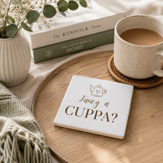 Cup of coffee on a square coaster with 'Fancy a Cuppa?' text, next to books and a vase with flowers.
