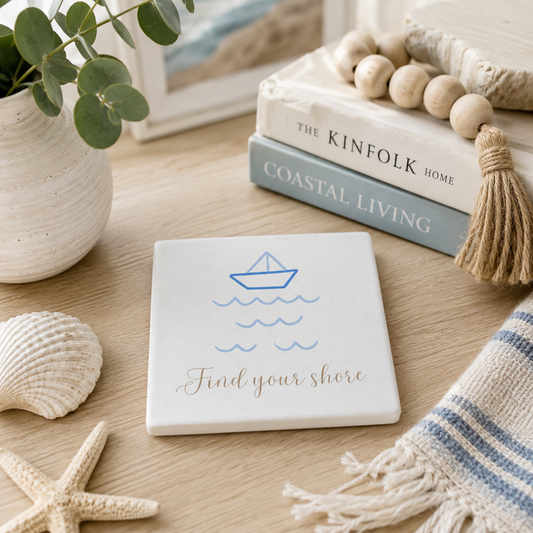 Square ceramic coaster with boat illustration and 'Find your shore' quote in blue and gold. The coaster is in a coastal theme room with shells and books.