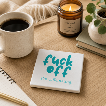 Square ceramic coaster with 'fuck off I'm caffeinating' text on a table with coffee mug, candle, and plant.