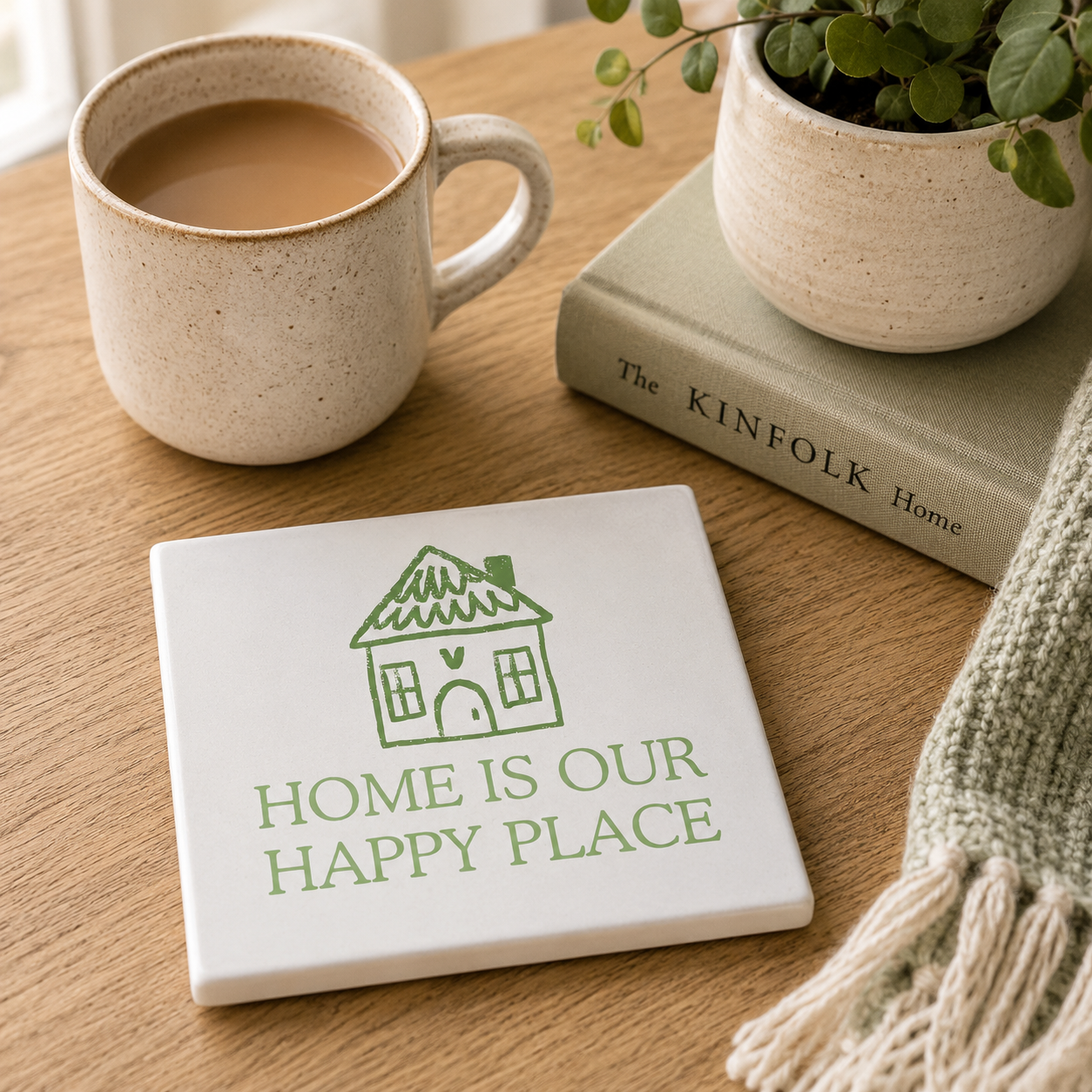 Square ceramic coaster with the quote 'Home is our happy place' and a cute house illustration. The coaster is set on a wooden surface with books and a cup of tea.