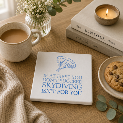 Square ceramic coaster featuring the funny quote "If at first you don't succeed, skydiving isn't for you" in crisp blue lettering with a parachute illustration, on a wooden table with tea and biscuits.