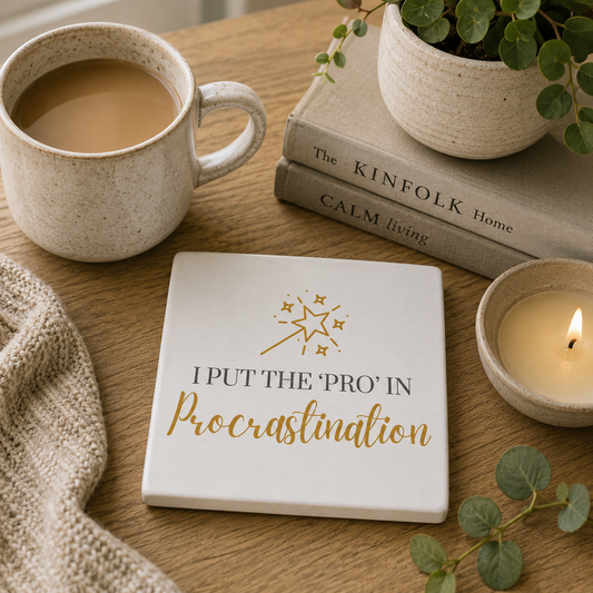 Square ceramic coaster featuring the funny quote "I put the 'pro' in procrastination" in crisp black and gold lettering with a magic wand and stars illustration, on a wooden surface with tea cup and books.
