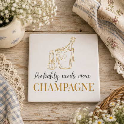Square ceramic coaster featuring the funny quote "Probably needs more champagne" in crisp grey and gold lettering with a champagne bottle illustration, on a wooden table.