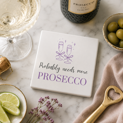 Square ceramic coaster featuring the funny quote "Probably needs more prosecco" in crisp grey and purple lettering with a prosecco glass illustration, on a marble surface with prosecco bottle and glasses.