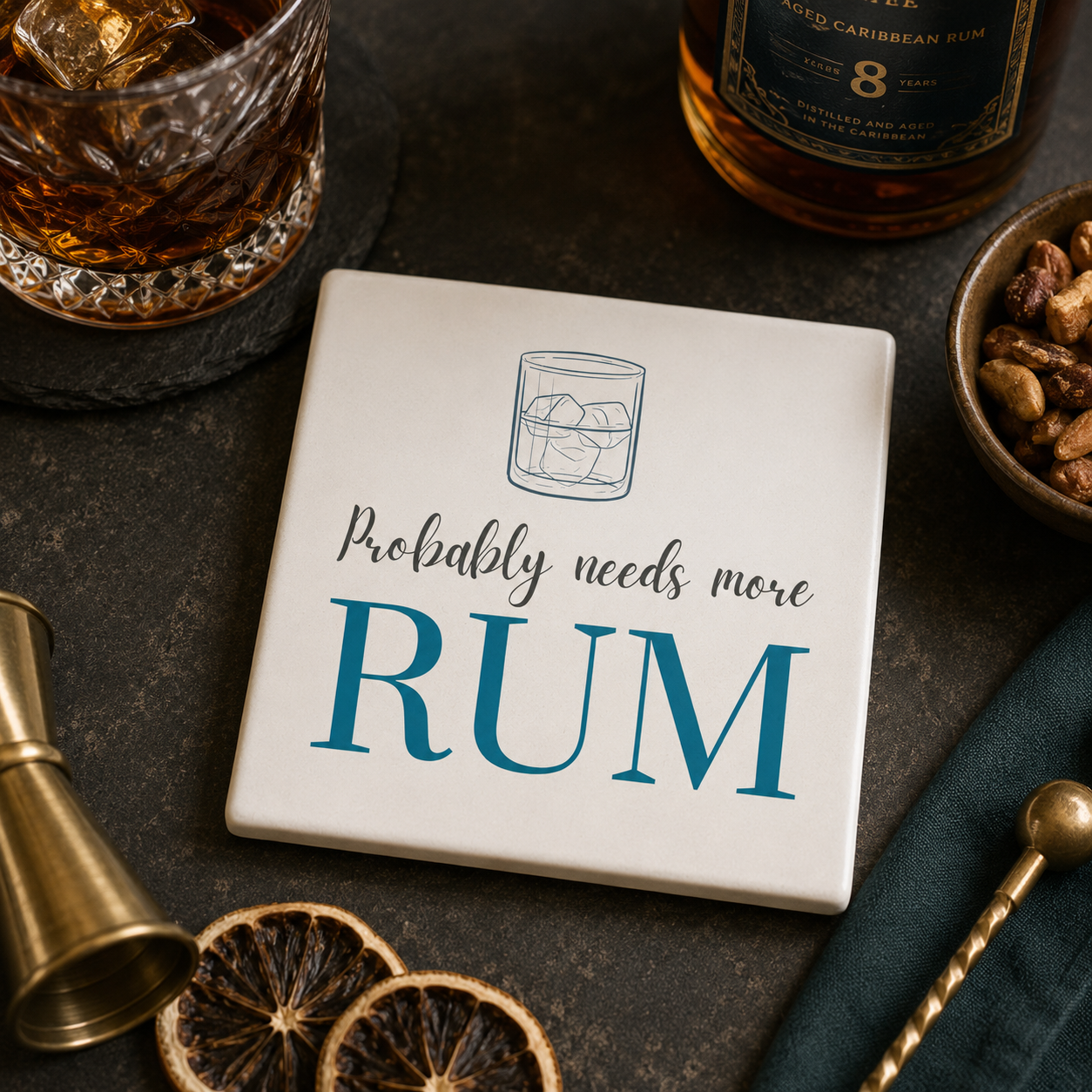 Square ceramic coaster featuring the funny quote "Probably needs more rum" in crisp grey and blue lettering with a rum glass illustration, on a bar with cocktail making accessories.