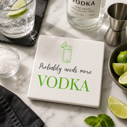 Square ceramic coaster featuring the funny quote "Probably needs more vodka" in crisp grey and green lettering with a vodka tonic glass illustration, on a marble surface with vodka cocktail making accessories.