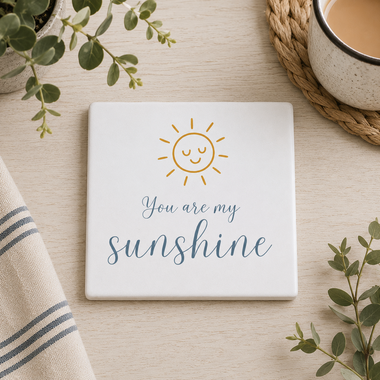 Square ceramic coaster featuring the quote "You are my sunshine" in blue lettering with a cute golden sun illustration, on a wooden surface with a cup of tea.
