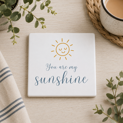 Square ceramic coaster featuring the quote "You are my sunshine" in blue lettering with a cute golden sun illustration, on a wooden surface with a cup of tea.