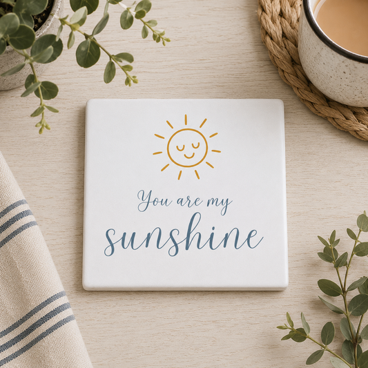 Square ceramic coaster featuring the quote "You are my sunshine" in blue lettering with a cute golden sun illustration, on a wooden surface with a cup of tea.