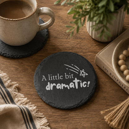 Round slate coaster with 'A little bit dramatic!' text on a wooden table with a cup of coffee and plant.
