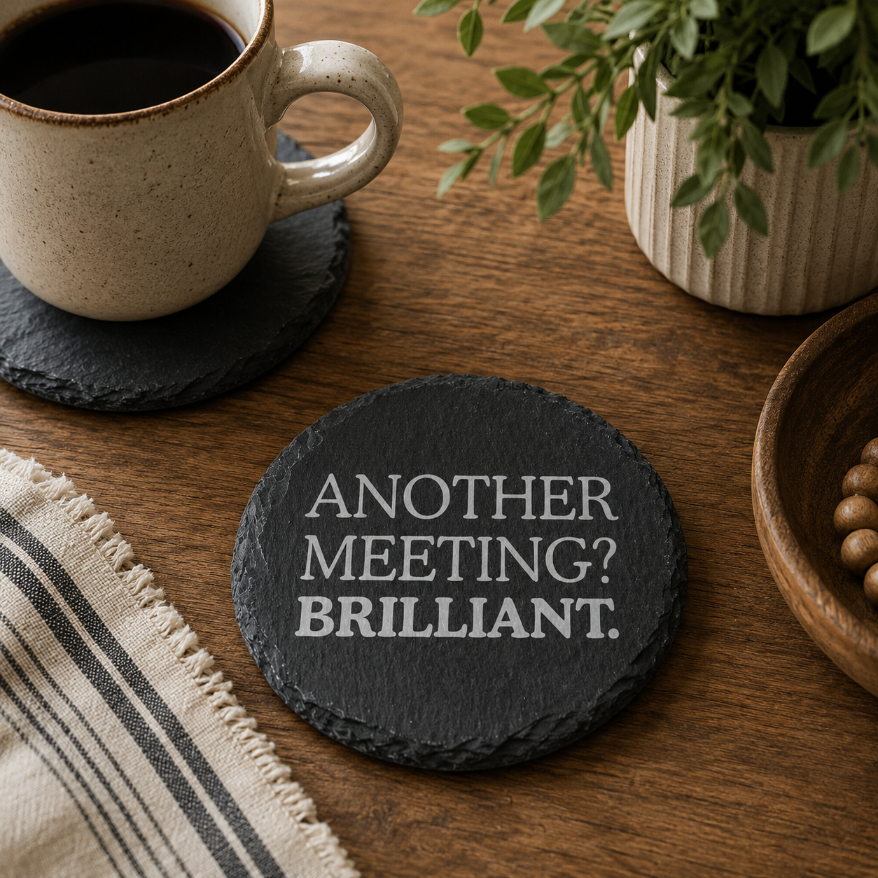 Round slate coaster with 'Another meeting? Brilliant.' text on a wooden table with a cup of coffee and plant.
