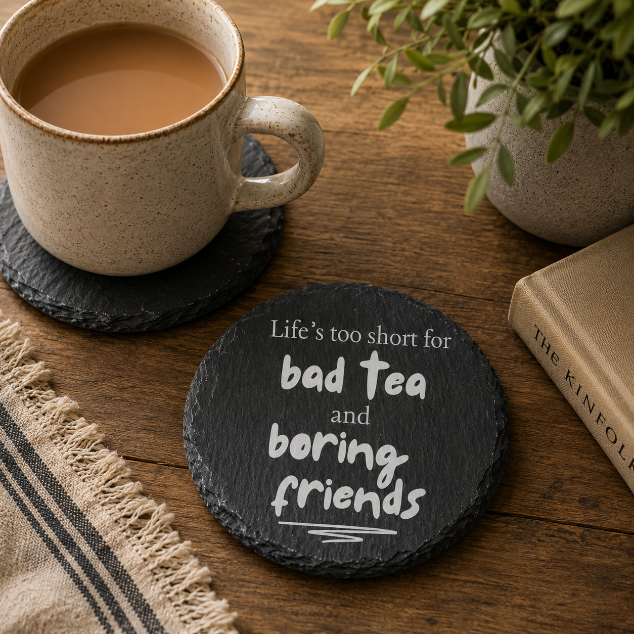 Round slate coaster with the quote 'life's too short for bad tea and boring friends' on a wooden table with mug of tea and books.