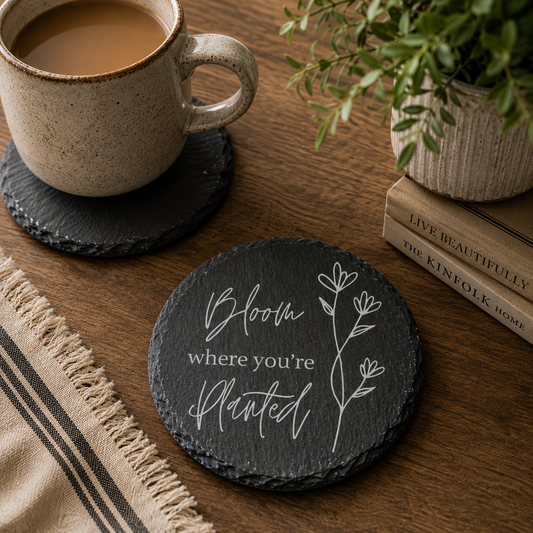Round slate coaster with 'Bloom where you're planted' text on a wooden coffee table with a cup of tea and books.