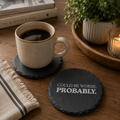 Round slate coaster reading 'Could be worse, probably.' on a wooden desk with cup of coffee.