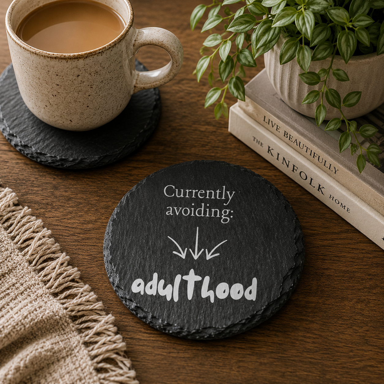 Round slate coaster with 'Currently avoiding: adulthood' text on a wooden table with a cup of coffee and books.
