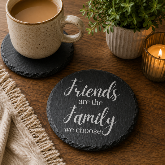 Round slate coaster with 'Friends are the family we choose' text on a wooden table with a cup of coffee and plant.