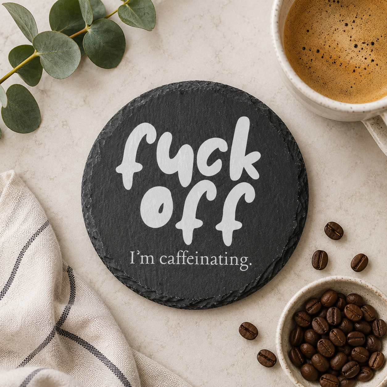 Round slate coaster with 'fuck off I'm caffeinating' text on a table with coffee and beans.
