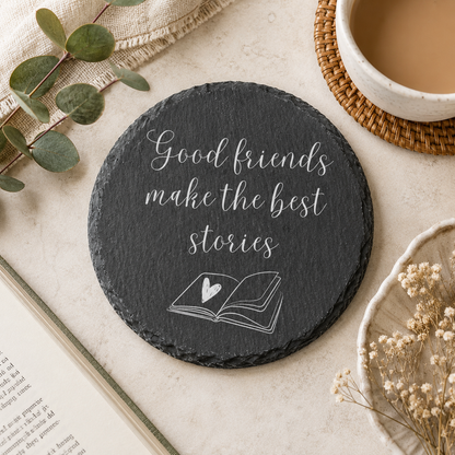 Round slate coaster with 'Good friends make the best stories' text on a table with a cup of coffee and book.