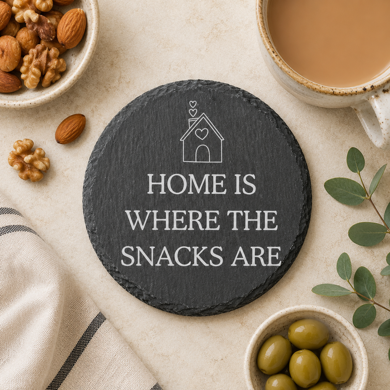 Round slate coaster with 'Home is where the snacks are' text, surrounded by snacks and a cup of coffee.