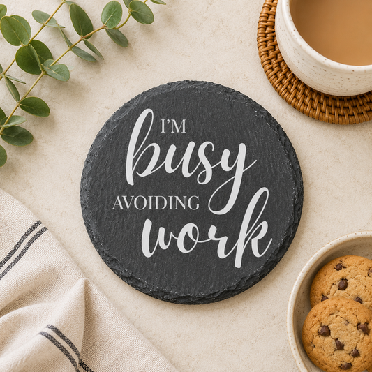 Round slate coaster with 'I'm busy avoiding work' text on a table with a cup, cookies, and plant.