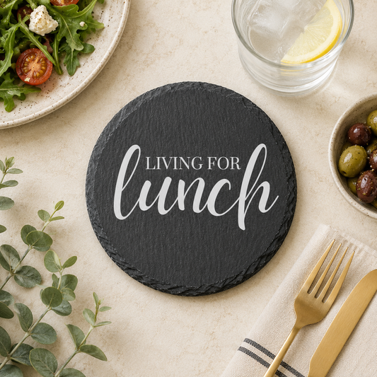 Round slate coaster with 'Living for Lunch' text on a table setting with salad, olives, and cutlery.