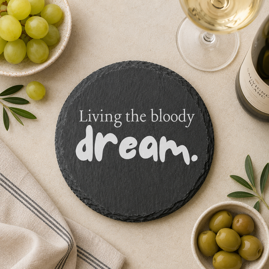 Round slate coaster with 'Living the bloody dream' text on a table with grapes, olives, and wine.