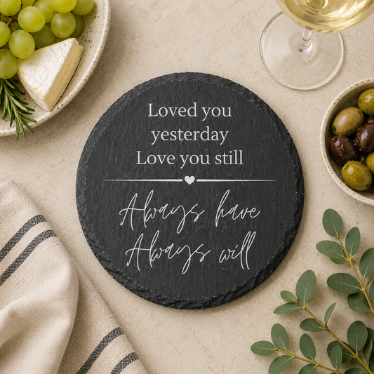 Round slate coaster with romantic quote on a table with cheese, grapes, and olives.