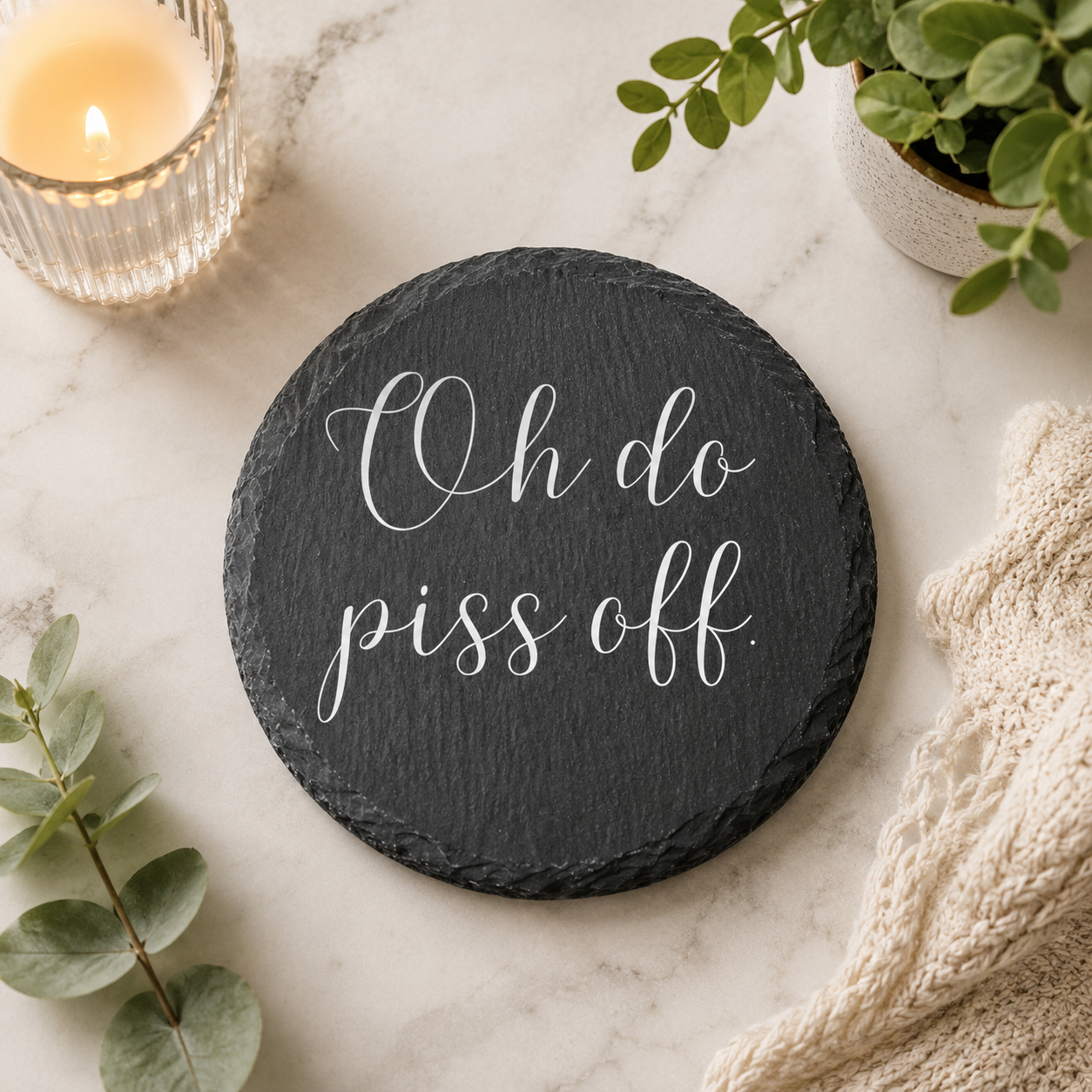 Round slate coaster with 'oh do piss off' text on a marble surface with candle and plants.