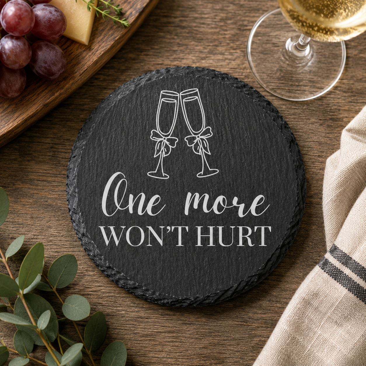 Round slate coaster with 'One more WON'T HURT' text and wine glass illustration on a wooden table with grapes and a glass of wine.