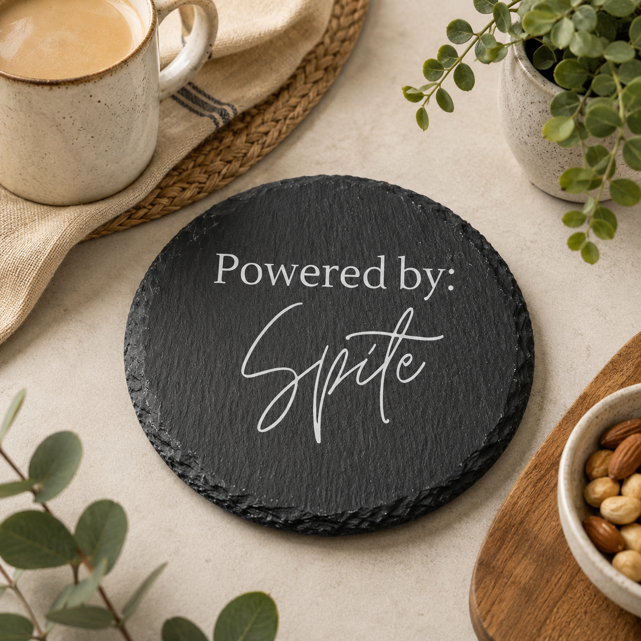 Round slate coaster with 'Powered by Spite' text on a table with coffee and snacks