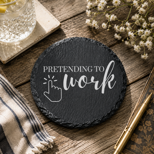 Round black slate coaster with 'Pretending to work' text on a wooden surface with flowers and a glass.