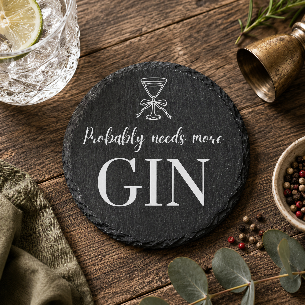 Round slate coaster with 'Probably needs more Gin' text on a wooden surface with a glass of gin and tonic.