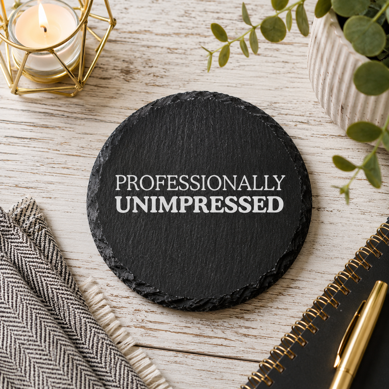 Round black slate coaster with 'PROFESSIONALLY UNIMPRESSED' text on a light wooden desk with office stationery.
