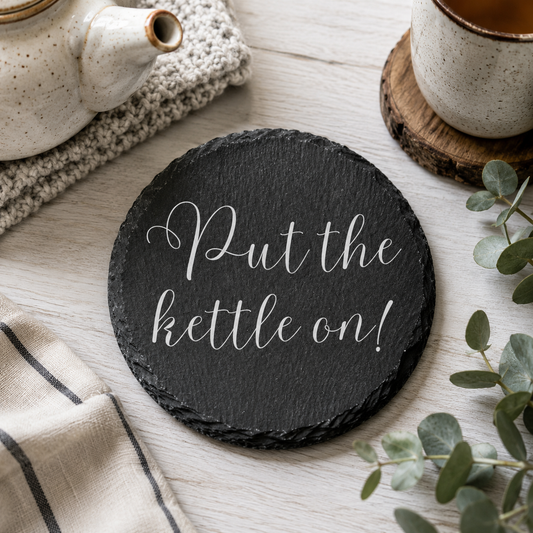 Round slate coaster with 'Put the kettle on!' text on a wooden surface with tea accessories.