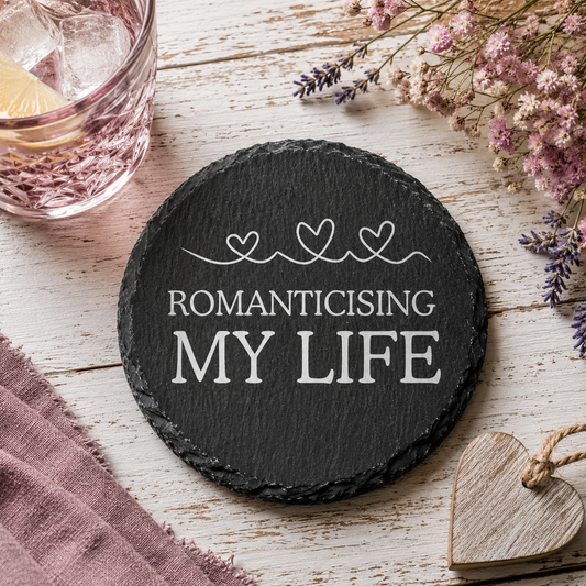 Round black slate coaster with 'ROMANTICISING MY LIFE' text on a wooden surface with flowers and a glass.