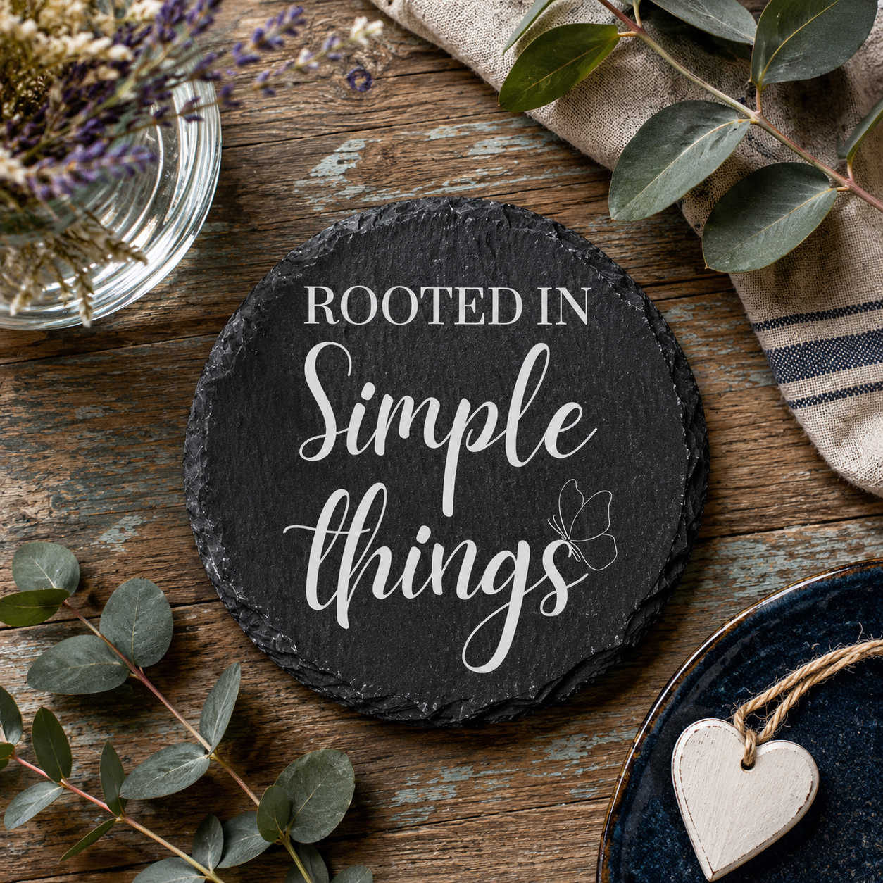 Round slate coaster with 'Rooted in Simple Things' text on a wooden surface with greenery and a heart-shaped object.