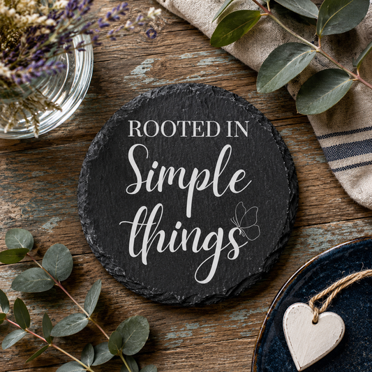 Round slate coaster with 'Rooted in Simple Things' text on a wooden surface with greenery and a heart-shaped object.