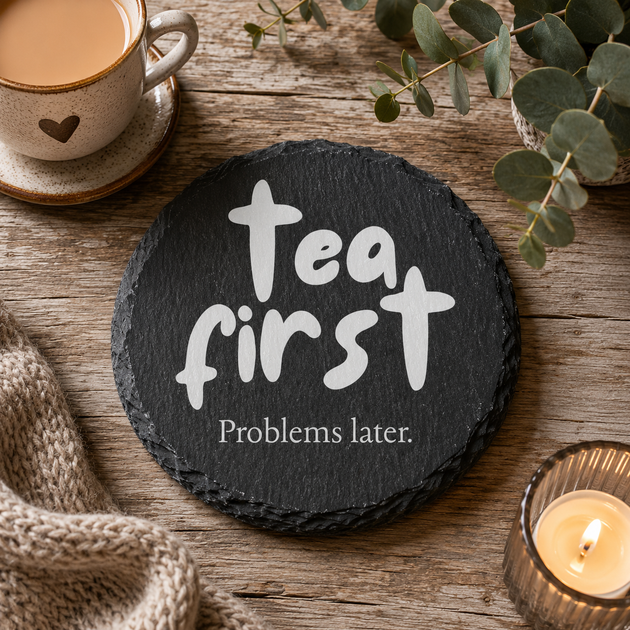 Round slate coaster with 'Tea first, Problems later' text on a wooden surface with a cup of tea, candle, and eucalyptus leaves.