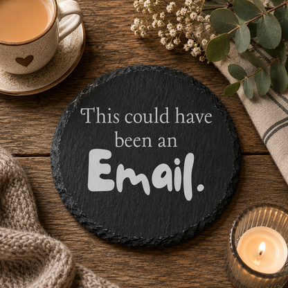 Round black slate coaster with text 'This could have been an Email' on a wooden table with a cup of coffee, candle, and flowers.