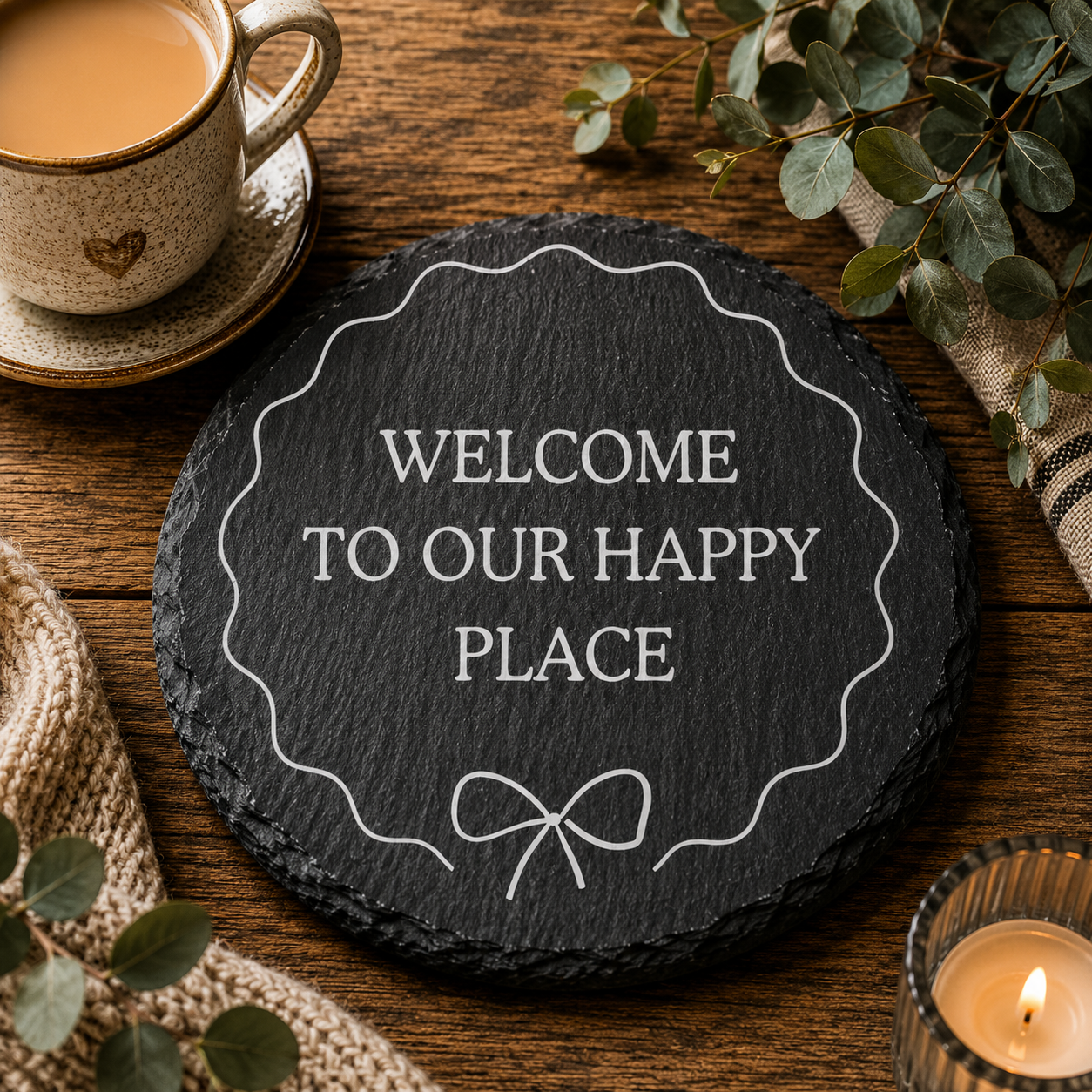Round slate coaster with 'Welcome to our happy place' text on a wooden table with a cup of tea and candle.
