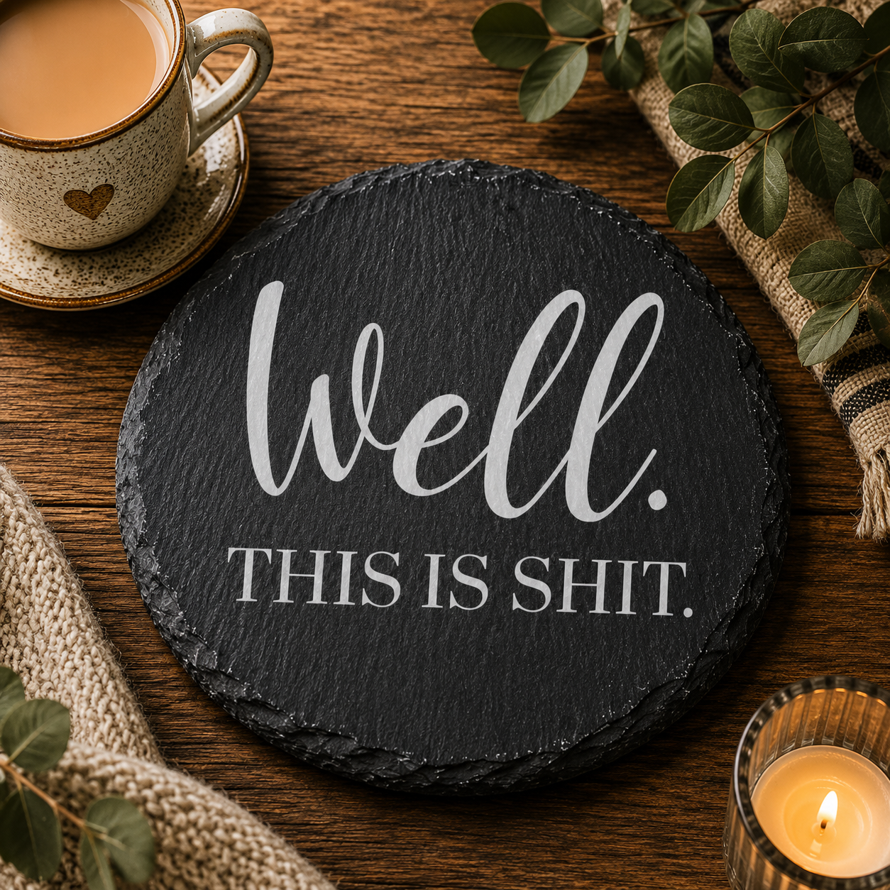 Round slate coaster with 'Well. This is shit.' text on a wooden table with a cup of coffee and candle.