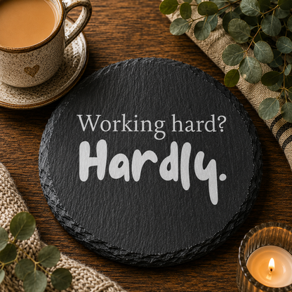 Round slate coaster with 'Working hard? Hardly.' text on a wooden table with a cup of coffee and decorative elements.