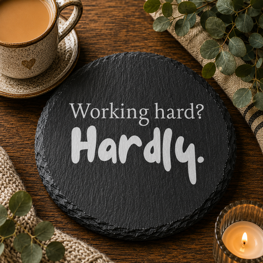 Round slate coaster with 'Working hard? Hardly.' text on a wooden table with a cup of coffee and decorative elements.