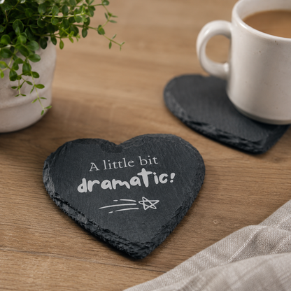 Heart-shaped slate coaster with "A little bit dramatic!" quote on a wooden table with a cup of coffee.