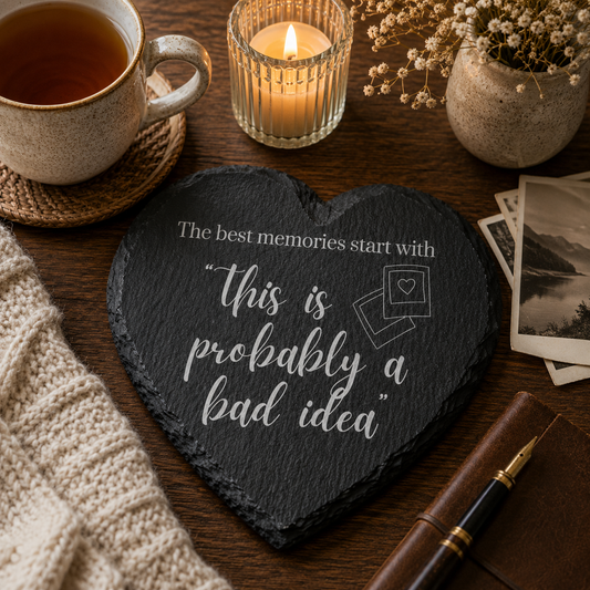 Heart-shaped slate coaster with humorous quote, mug with tea, candle, and photos on a wooden table.
