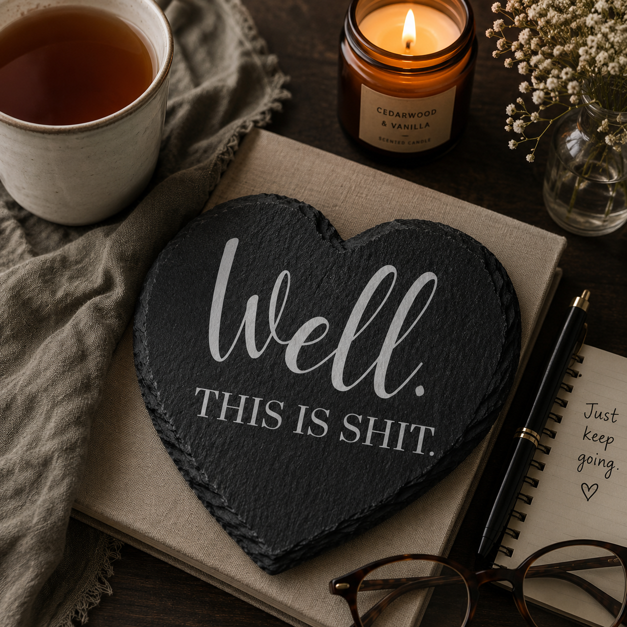 Heart-shaped slate coaster with 'Well. This is shit.' text, cup of tea, candle, and notebook on a wooden surface.