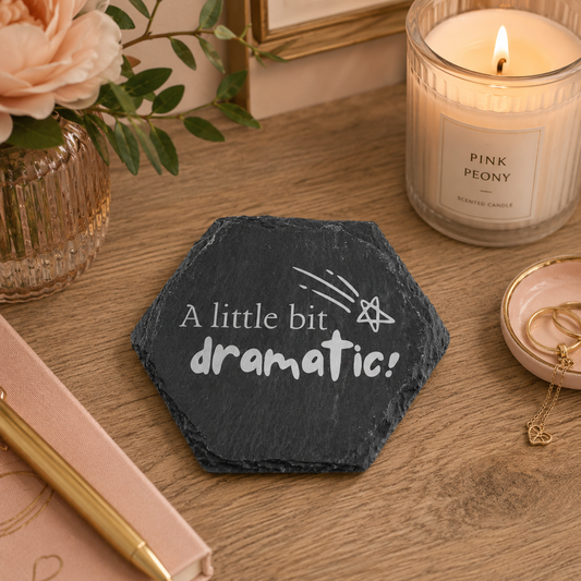 Hexagonal slate coaster with 'a little bit dramatic!' text on a wooden surface with flowers, candles and jewellery.